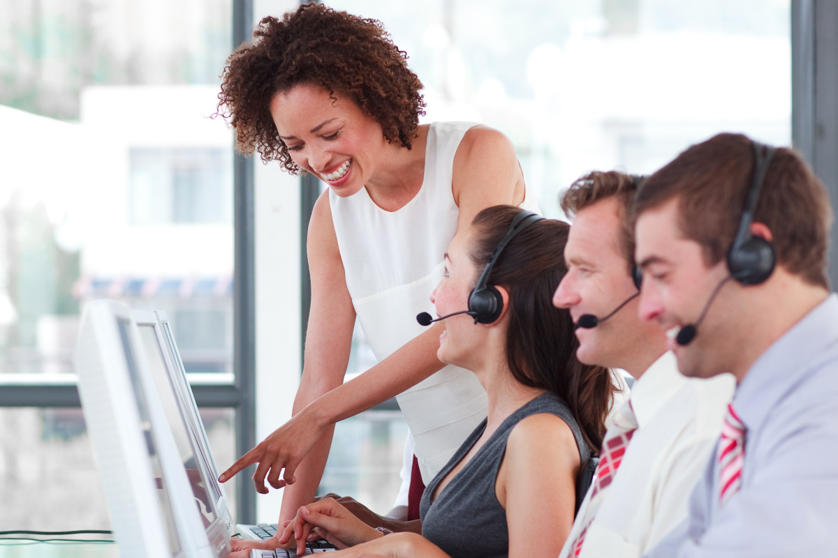 A group of workers with headsets taking calls with a smiling supervisor assisting them